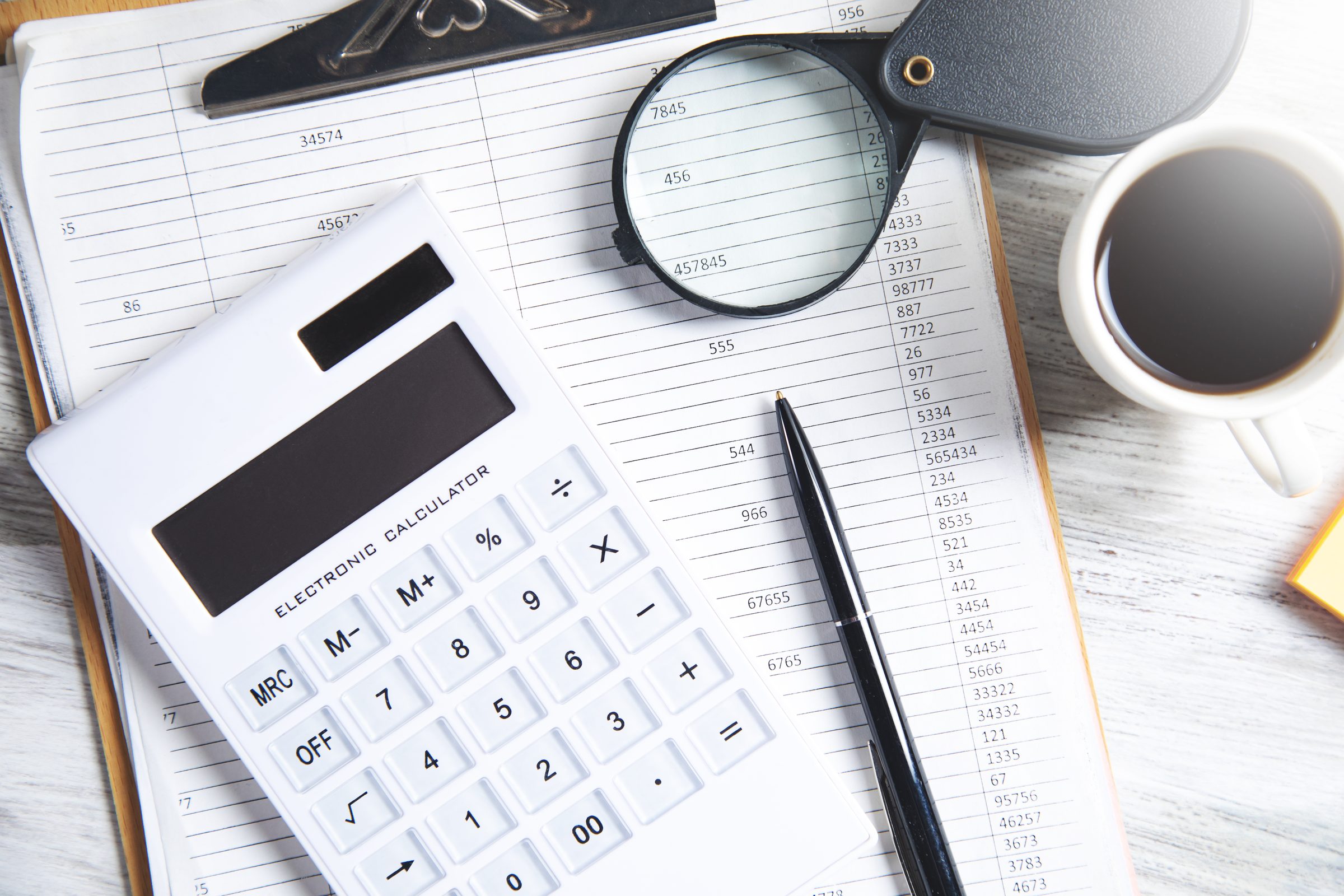 A calculator, a ledger, a magnifying glass and coffee on a desk